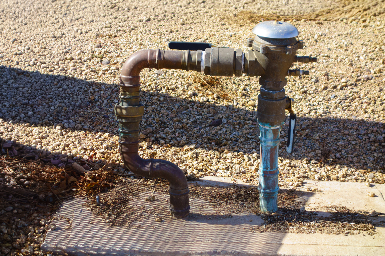 outdoor-brass-pressure-vacuum-breaker-backflow-preventer-assembly-unit-connected-to-weathered-copper-water-plumbing-pipe-lines-over-gravel-ground-on-sunny-day